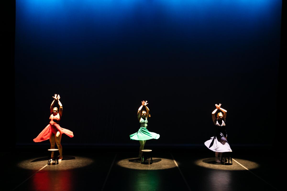 3 dancers in motion, red, green, and white skirts, respectively in pools of light, skirts fanned out from movement or twirling, arms raised and crossed at the wrists