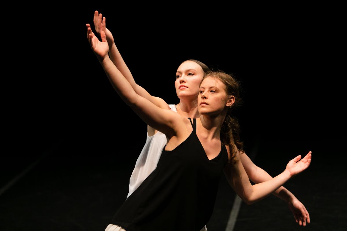 2 dancers, one behind the other, with right arm stretched out and upward and left stretched out and down