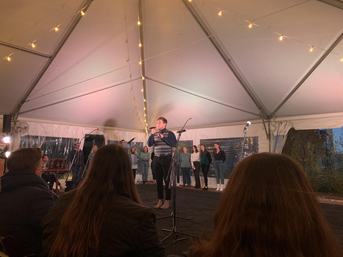 Person with short hair speaking into a microphone to a seated audience under a lighted tent.